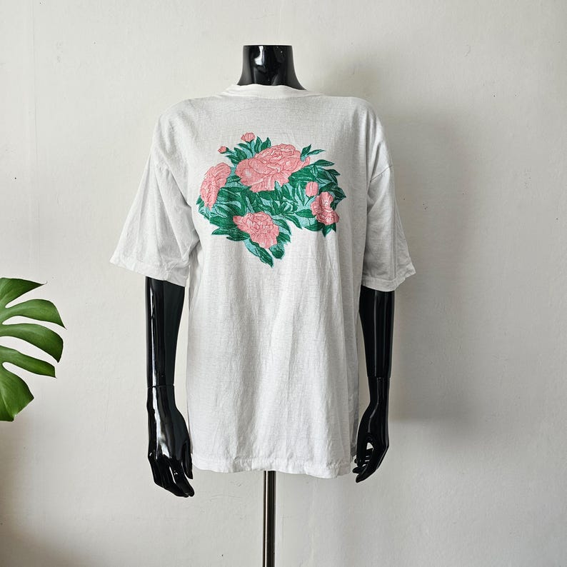 Op de afbeelding: Een lichtgrijs t-shirt met een bloemenontwerp. De grafische print toont roze bloemen en groene bladeren. Het t-shirt wordt getoond op een zwarte mannequin tegen een witte achtergrond.