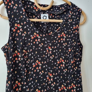 K&ouml;nnte beinhalten: Ein &auml;rmelloses schwarzes Kleid mit einem kleinen Blumenmuster in Orange-, Gelb- und Lilat&ouml;nen. Das Kleid hat einen V-Ausschnitt und einen taillierten Oberteil.