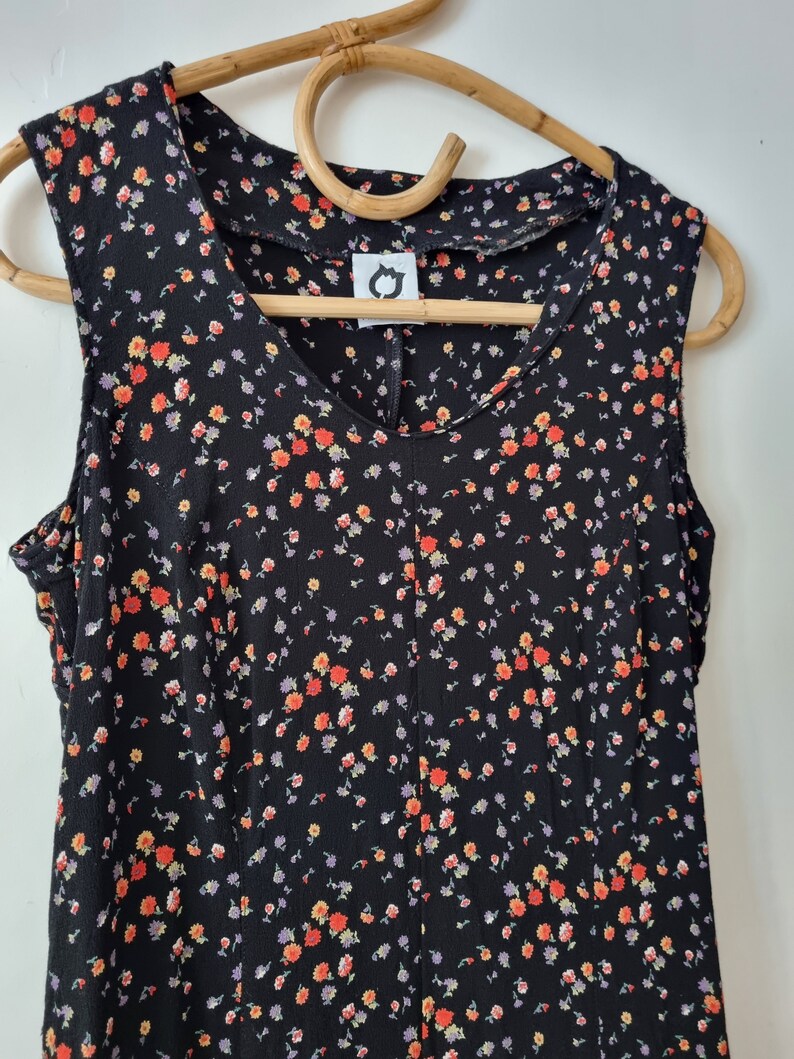 K&ouml;nnte beinhalten: Ein &auml;rmelloses schwarzes Kleid mit einem kleinen Blumenmuster in Orange-, Gelb- und Lilat&ouml;nen. Das Kleid hat einen V-Ausschnitt und einen taillierten Oberteil.