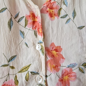May include: A white button-up shirt with a floral print featuring pink, orange, and yellow flowers with green leaves. The shirt has a V-neck and three white buttons.