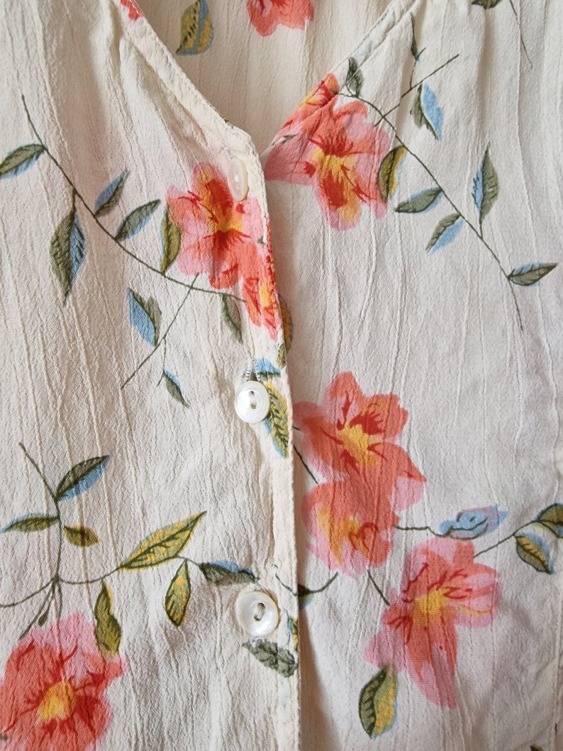 May include: A white button-up shirt with a floral print featuring pink, orange, and yellow flowers with green leaves. The shirt has a V-neck and three white buttons.
