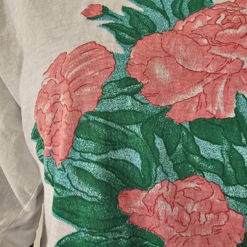Op de afbeelding: Close-up van een lichtgrijs shirt met lange mouwen met een bloemenontwerp. Het ontwerp bevat grote roze bloemen met groene bladeren en een lichtblauwe achtergrond. De stof van het shirt lijkt zacht en comfortabel.