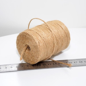 Peut inclure: Une bobine de ficelle marron naturel est posée à côté d'une règle métallique. La ficelle est bien enroulée et possède une boucle à une extrémité. La règle affiche des mesures en centimètres.