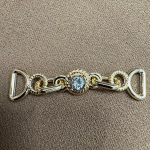 May include: Gold-toned metal buckle with a clear rhinestone in the center. The buckle has a chain-like design and two D-shaped loops on either end.