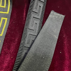 Puede incluir: Primer plano de cinta decorativa con patrones geom&eacute;tricos. La cinta presenta un dise&ntilde;o de clave griega en negro y gris, con un patr&oacute;n dorado y negro visible a la izquierda. El fondo es de terciopelo rojo oscuro.
