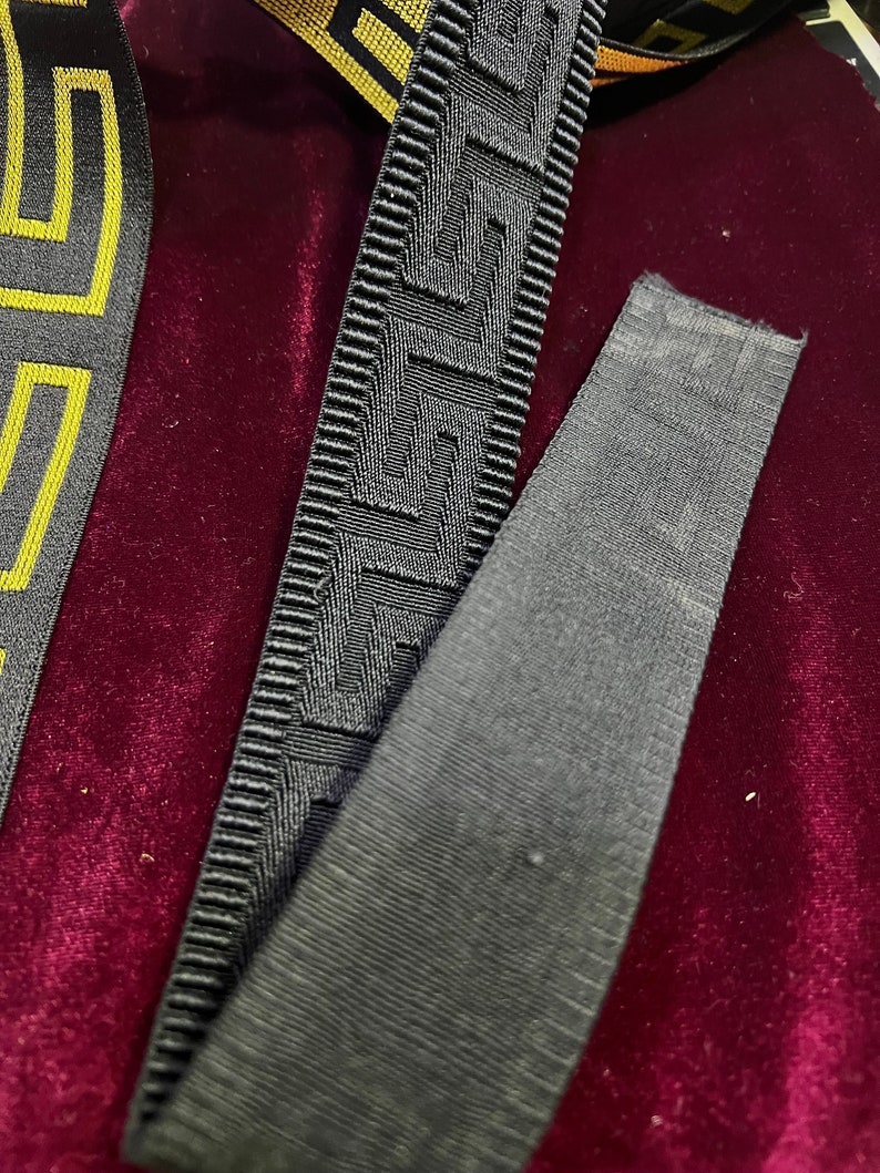 Puede incluir: Primer plano de cinta decorativa con patrones geom&eacute;tricos. La cinta presenta un dise&ntilde;o de clave griega en negro y gris, con un patr&oacute;n dorado y negro visible a la izquierda. El fondo es de terciopelo rojo oscuro.