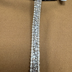May include: Silver rhinestone trim featuring a row of larger rhinestones and a row of smaller rhinestones. The trim is displayed on a brown fabric background.