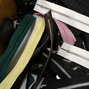 May include: A selection of assorted zippers in colours such as black, white, green, and pink. The zippers feature metal teeth and vary in length. Some zippers have gold-coloured teeth.