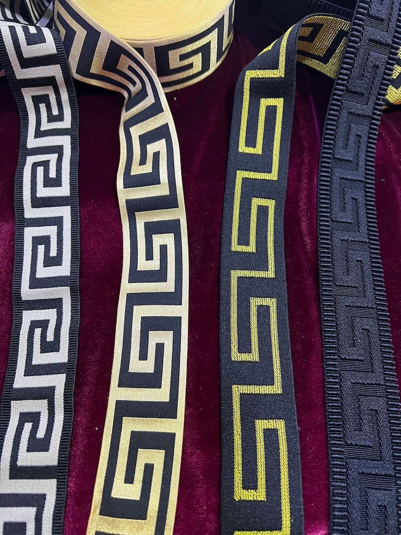 Puede incluir: Primer plano de rollos de cintas decorativas en varios colores. Las cintas presentan un patr&oacute;n de greca en negro, dorado y plateado. Las cintas est&aacute;n hechas de un material tejido y son adecuadas para manualidades y adornos.