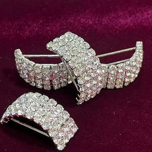Può includere: Tre fermagli a barretta con strass argentati. Ogni fermaglio presenta più file di strass trasparenti e scintillanti in un design curvo. I fermagli sono esposti su una superficie di velluto bordeaux scuro, creando un contrasto.