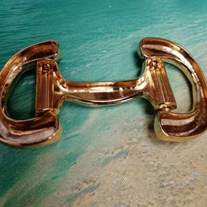 May include: A gold-colored metal horse bit, a type of equestrian equipment. The bit has a curved shape with rounded ends and a central bar. The metal has a shiny, reflective surface.