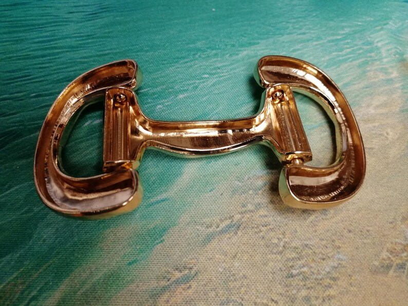 May include: A gold-colored metal horse bit, a type of equestrian equipment. The bit has a curved shape with rounded ends and a central bar. The metal has a shiny, reflective surface.