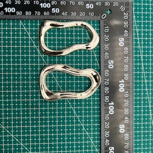 May include: Two silver-colored, abstract-shaped metal objects. Each is approximately 4 inches long, with a curved, open design. They are placed on a green grid surface with a ruler for scale.