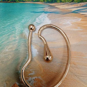 May include: A gold-colored ear cuff with a curved design. The cuff features a small ball at the top and a teardrop-shaped element. The jewelry is set against a beach-themed background.