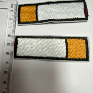 May include: Two embroidered iron-on patches shaped like cigarettes. The patches are white with a black outline and a yellow and red detail. The patches are approximately 2 inches long.