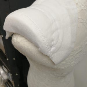 Puede incluir: Una hombrera blanca curvada hecha de un material suave similar al fieltro. El accesorio presenta líneas de costura paralelas visibles a lo largo de su borde redondeado, diseñadas para dar estructura y forma al área del hombro de una prenda.