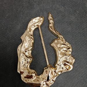 Pode incluir: Broche dourado de formato abstrato com superfície texturizada. O broche tem um alfinete na parte de trás para fixação. O design é orgânico e fluido, com aparência de metal derretido.