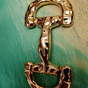 May include: A shiny gold-colored metal horse bit, featuring a curved top and bottom with a central vertical bar. The bit has a reflective surface, and is set against a green and blue background.