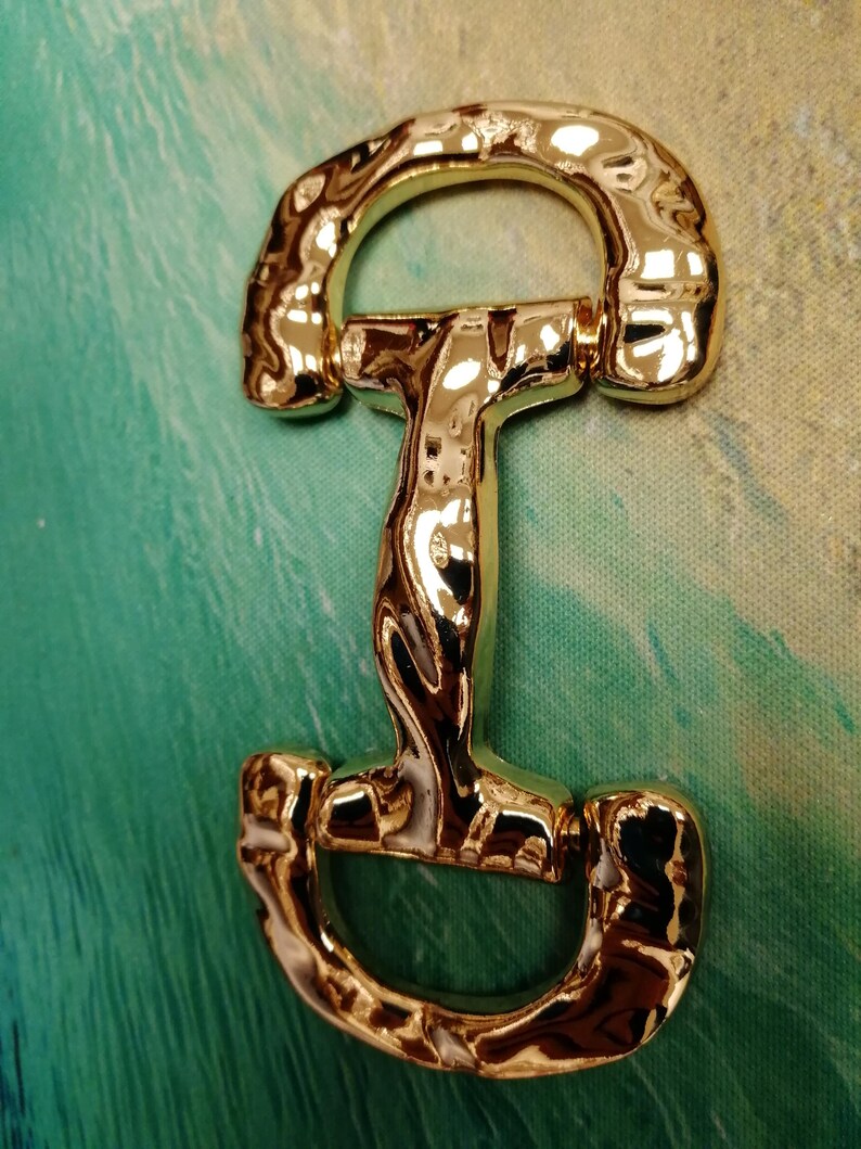 May include: A shiny gold-colored metal horse bit, featuring a curved top and bottom with a central vertical bar. The bit has a reflective surface, and is set against a green and blue background.