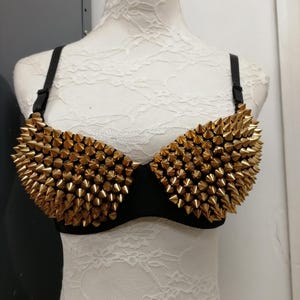 May include: Black bra with gold-colored cone studs covering the cups. The bra has black straps and is displayed on a white mannequin. The studs are densely packed, creating a bold, edgy look. The bra is a unique fashion statement.