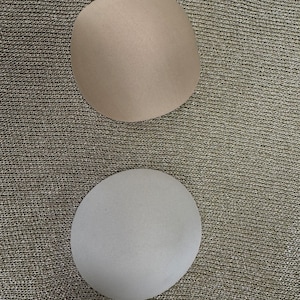 Puede incluir: Dos almohadillas para sujetador, una de color beige claro y otra de color gris claro. Ambas almohadillas tienen forma redondeada y una superficie texturizada. El fondo es una tela texturizada y metálica.