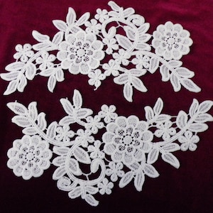 Pode incluir: Dois apliques de renda branca com desenhos florais. Cada aplique apresenta uma flor grande com flores menores e folhas.