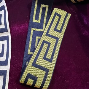 Puede incluir: Primer plano de cinta decorativa con un patr&oacute;n geom&eacute;trico de clave griega. La cinta presenta un fondo azul marino con un dise&ntilde;o dorado. La cinta se muestra sobre una superficie de terciopelo morado oscuro.