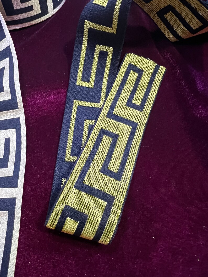 Puede incluir: Primer plano de cinta decorativa con un patr&oacute;n geom&eacute;trico de clave griega. La cinta presenta un fondo azul marino con un dise&ntilde;o dorado. La cinta se muestra sobre una superficie de terciopelo morado oscuro.