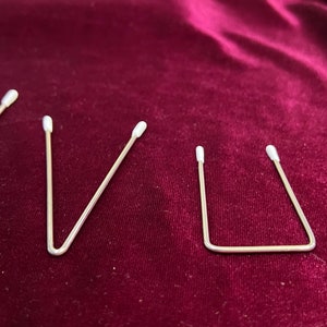 May include: Four silver metal wire hair clips with white plastic tips. The clips are arranged in a row on a dark red velvet surface.