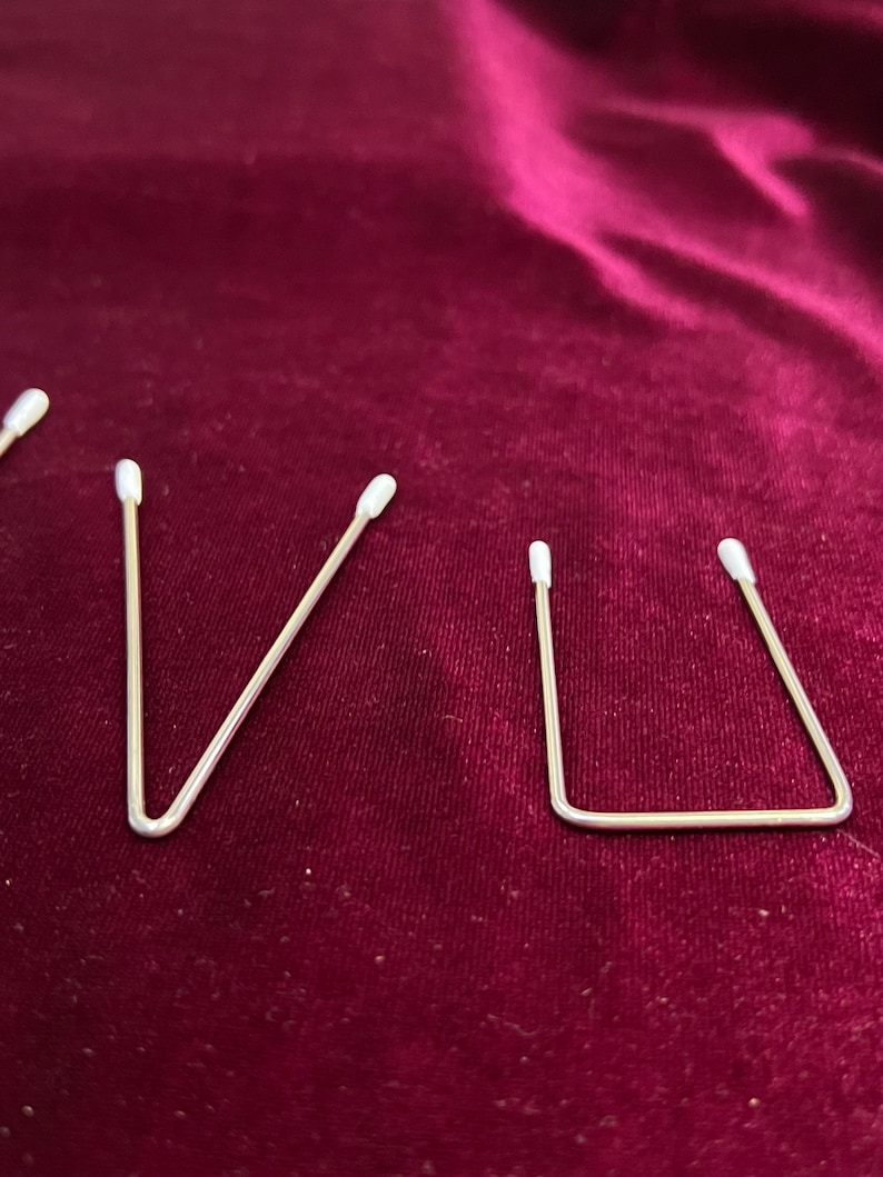 May include: Four silver metal wire hair clips with white plastic tips. The clips are arranged in a row on a dark red velvet surface.