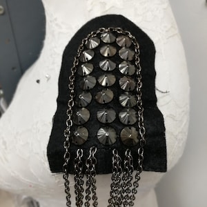 May include: A black shoulder embellishment with rows of silver-toned, faceted studs and dangling chain fringe. The accessory is attached to a black felt base and is displayed on a white mannequin form, showcasing a punk rock aesthetic.