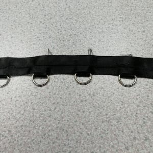 May include: A black fabric ribbon strip featuring four evenly spaced silver-toned metal D-rings attached along the bottom edge. The ribbon has a smooth, woven texture with visible stitching securing the metal hardware in place along the length.