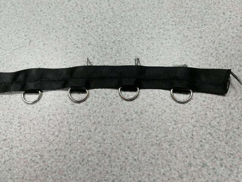 May include: A black fabric ribbon strip featuring four evenly spaced silver-toned metal D-rings attached along the bottom edge. The ribbon has a smooth, woven texture with visible stitching securing the metal hardware in place along the length.
