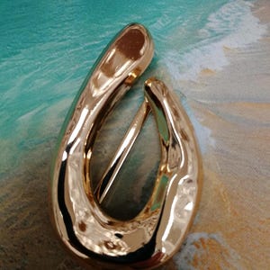 Könnte beinhalten: Ein goldfarbener, tropfenförmiger Haarklip mit glänzender, reflektierender Oberfläche. Der Clip hat ein schlichtes, elegantes Design und einen Bügel, der das Haar fixiert. Der Hintergrund ist eine Strandszene.