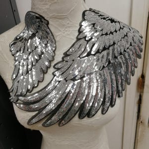 Puede incluir: Adornos en forma de alas con lentejuelas plateadas. Las alas están hechas de numerosas lentejuelas pequeñas y brillantes adheridas a un respaldo negro. Se exhiben en un maniquí blanco, mostrando su forma y diseño. Un artículo decorativo.