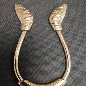 May include: A gold-colored curtain tieback with a textured, leaf-like design at each end. The tieback has a curved, U-shaped body and is made of a shiny, metallic material. The overall design is elegant and decorative.