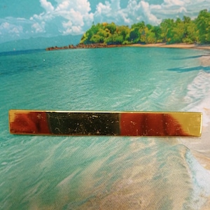 May include: A rectangular gold-toned tie bar with a central section of reddish-brown color. The tie clip is a classic accessory for formal wear, adding a touch of elegance to any outfit. The bar is approximately 2 inches long.