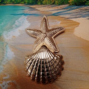 Könnte beinhalten: Ein goldfarbenes dekoratives Element in Form eines Seesterns und einer Muschel. Der Seestern ist über der Muschel positioniert und erzeugt ein Küsten- oder maritimes Thema. Der Artikel ist wahrscheinlich ein Dekorationsobjekt.