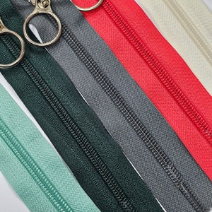 May include: Five different colours of zipper tape with metal zipper pulls. The colours are green, dark green, grey, red, and cream.