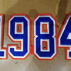 May include: Embroidered patch displaying the year "1984". The blue numbers have a white border and an orange outline. This patch could be for a jacket or bag.