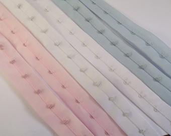 Könnte beinhalten: Fünf Streifen BH-Band mit Haken- und Ösenverschluss in Rosa, Weiß und Hellblau. Das Band ist aus elastischem Material und hat Reihen von Metallhaken und Ösen.
