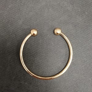 May include: A gold-coloured open cuff bracelet. The bracelet features a smooth, rounded band with a small, spherical accent at each open end. The jewellery is displayed on a dark grey surface.