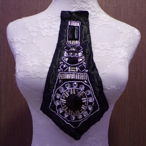 May include: A black fabric neck piece with detailed embellishments. It features a central column of rectangular and oval black stones, surrounded by silver beads and clear crystals. The piece is displayed on a white mannequin.