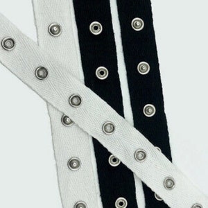 Puede incluir: Dos cintas de adorno blancas y negras con ojales plateados. La cinta blanca tiene un patrón de espiga.