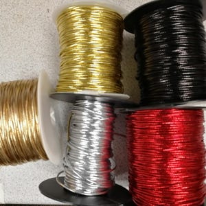 Könnte beinhalten: Fünf Spulen mit metallischer Kordel in Gold, Schwarz, Silber und Rot. Die Spulen sind mit einer goldenen Spule auf der linken Seite und einem Stapel von vier Spulen in Gold, Schwarz, Silber und Rot auf der rechten Seite angeordnet. Jede Spule ist mit einer dünnen, glänzenden Kordel umwickelt.