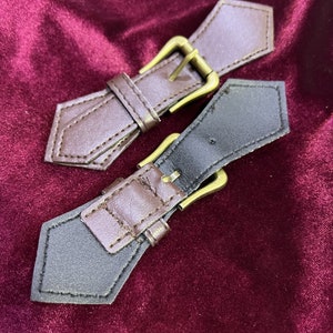 Puede incluir: Dos hebillas de cinturón de cuero marrón con cierres de metal dorado. Las hebillas son puntiagudas en los extremos y tienen costuras alrededor de los bordes.