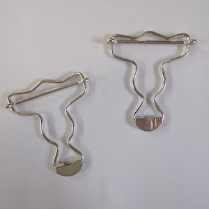 Könnte beinhalten: Zwei silberne Metall-Kleidungsverschlüsse mit wellenförmigem Design. Die Verschlüsse sind zum Befestigen von Kleidungsstücken gedacht.