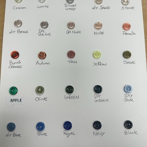 Puede incluir: Una colección de botones redondos en varios colores, incluyendo crema, blanco y gris plateado. Los botones están dispuestos sobre una superficie blanca, con cada color etiquetado. Otros colores incluyen beige, nude, melocotón, naranja, amarillo, verde, azul y negro.