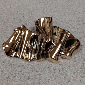 May include: A gold-coloured, abstract metal brooch with a flowing, ribbon-like design. The brooch has a shiny, reflective surface and appears to be made of a solid metal. The design is sculptural and three-dimensional.