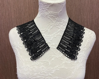 May include: Black lace collar with a scalloped edge, displayed on a white mannequin. The collar has a delicate, openwork design, and is a stylish accessory. The background is a brown, vertically striped pattern.